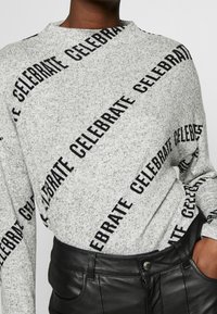 Grå, texturerad tröja med svart diagonalt mönster av texten "CELEBRATE" och rund halsringning. Sammanställd med högmidjade svarta läderbyxor.