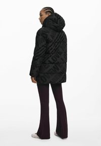 Doudoune noire avec capuche surdimensionnée, arborant un motif de texte en relief sur l'ensemble. Associée à un jean évasé foncé et des chaussures de couleur claire.