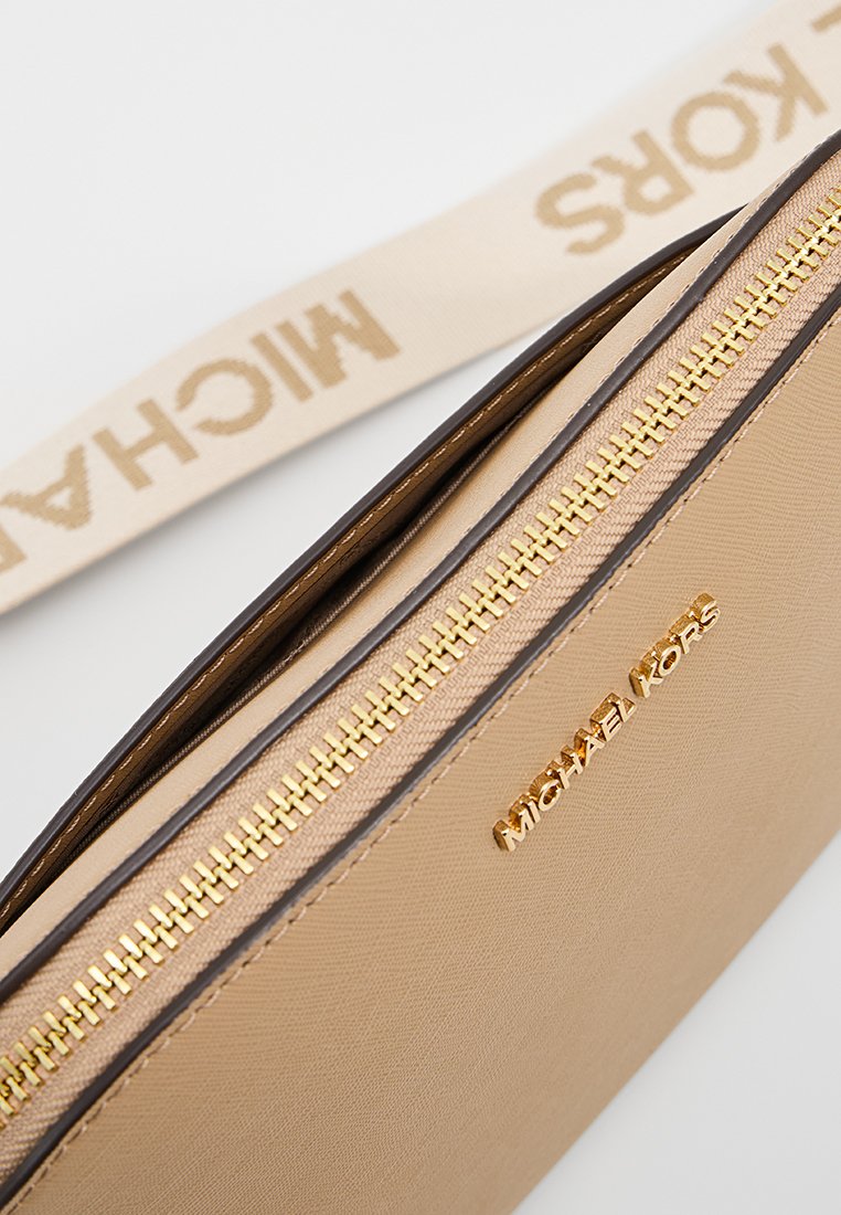 Pochette en cuir beige avec fermeture éclair et quincaillerie dorées. Présente une bandoulière beige avec le logo "MICHAEL KORS". Texture lisse et design minimaliste.