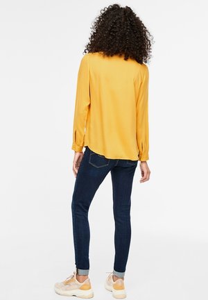 Femme aux cheveux bouclés portant un chemisier à manches longues jaune moutarde, un jean skinny bleu foncé et des baskets beiges, debout de dos.