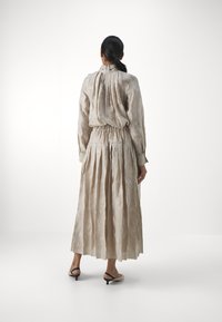 Rochie bej, cu model, cu guler înalt, mânecă lungă, talie elastică și fustă plisată; purtată cu mules negre cu toc jos.