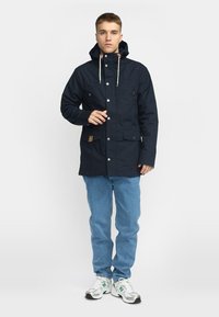 Navy waterproof jacket with a hood, snap buttons, and front pockets. Features a drawstring at the neck and brown leather accent on the pocket.