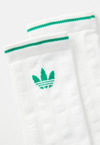 Baltie sporta zeķes ar zaļām svītrām augšdaļā un zaļu logotipu ar trim lapām. Izgatavotas no strukturēta auduma komfortam.