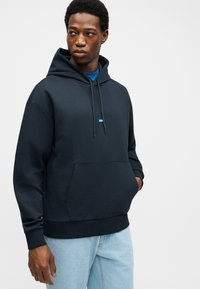 Νεαρός άνδρας που φοράει μαύρο φούτερ με κουκούλα και τσέπη στο μπροστινό μέρος, καθώς και ανοιχτό μπλε τζιν παντελόνι, κοιτάζει προς τα πλάγια με το ένα χέρι στην τσέπη.