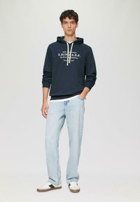Felpa navy con testo logo bianco, collo con cordini, abbinata a jeans in denim chiaro e sneakers bianche con una striscia nera.