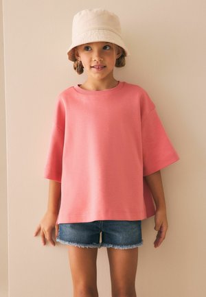 Tricou oversized de culoare roz deschis, cu mâneci scurte și guler rotund, asortat cu pantaloni scurți din denim cu margini ciufulite, pe un fundal neutru.