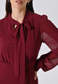 Burgunderfarbenes Kleid mit gebundenem Schleifen-Ausschnitt, transparenten langen Ärmeln und taillierter Passform. Sanfter Stoff mit subtiler Textur und eleganter Drapierung.