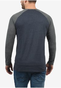 Mand iført en marineblå langærmet skjorte med grå ærmer og blå jeans, vist fra ryggen mod en hvid baggrund.