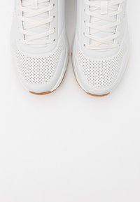 White athletic shoes with perforated design, rubber sole, and woven laces. The soles are brown, contrasting with the upper material.