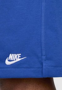 Blå shorts i tyg med mjuk textur, med en vit Nike-logotyp broderad på den nedre vänstra fållen och kontrasterande sömnadsdetaljer.