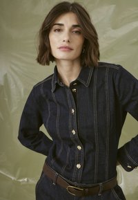 Chemise en denim sombre avec un design ajusté, des coutures bien visibles, des boutons dorés et une ceinture marron, sur un fond vert clair.