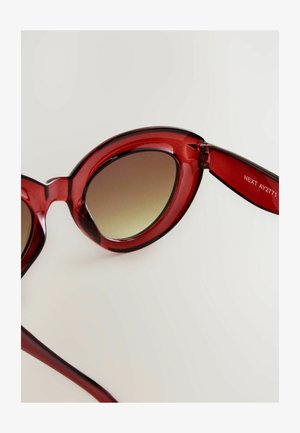 Røde, transparente katteøje-solbriller med gradient brune linser, en glat overflade og et slankt hængselsdesign. Mærket med "NEXT AY2771."