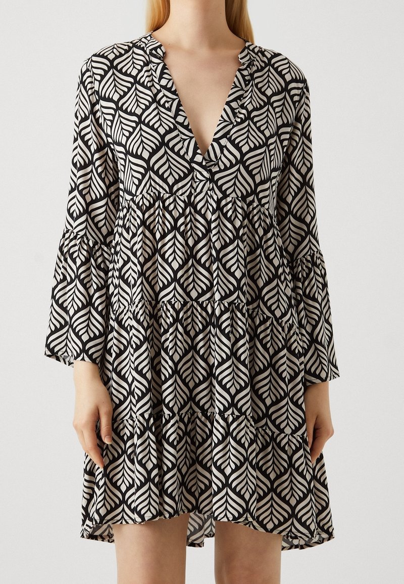 Robe noire et blanche à manches longues avec un grand motif de feuilles, un col en V profond, une jupe à étages et des manches évasées, portée par une personne à la peau claire et aux cheveux blonds.