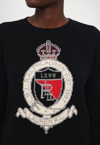 Zwarte gebreide trui met een opvallende trui patch in rood, wit en kastanjebruin, voorzien van een kroonembleem en tekstdetails.