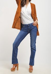 Bruine suède jas over een witte top, gecombineerd met blauwe rechttoe rechtaan jeans en tan platformhakken met houten blokken.