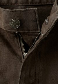 Primer plano de pantalones vaqueros marrones con cremallera abierta y un botón de metal con la etiqueta "KING DENIM" en la cintura.