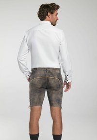 Bruine lederhosen met decoratieve stiksels en eventueel zijkoorden, gecombineerd met een aansluitend wit shirt met knoopmanchetten. Korte lengte.
