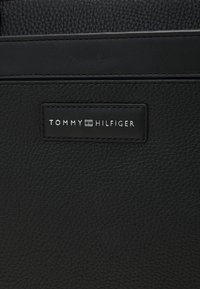 Sac à main en cuir noir avec une finition texturée. Présente une étiquette de marque rectangulaire portant l'inscription "TOMMY HILFIGER" en police argentée minimaliste.