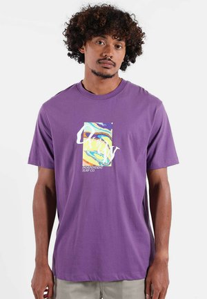 Homme aux cheveux bouclés portant un t-shirt violet avec un graphisme rectangulaire coloré et le texte blanc "Independent Surf Co." à l'avant.