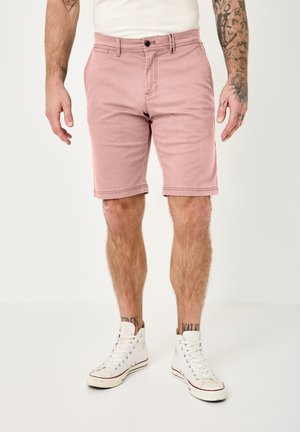 Man wearing light pink knee-length shorts, white high-top sneakers, and a white shirt, standing against a plain background.