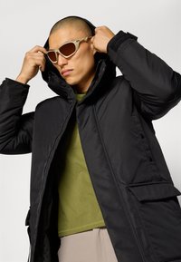 Parka negra e insulated con un cuello alto, cremallera frontal y bolsillos laterales, llevada sobre una camiseta verde. Acentuada con gafas de sol beige.