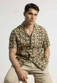 Camisa marrón de manga corta con patrones geométricos en color crema; diseño de botonadura y un ajuste relajado. El modelo lleva pantalones beige claros.
