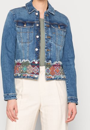 Denimjakke med sølvfarvede knapper og farverige broderede geometriske mønstre langs kanten, båret over en grøn top og cremefarvede bukser.