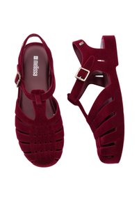 Sandalias de terciopelo rojo con la punta cerrada y el talón abierto, que presentan tiras entrelazadas y una hebilla ajustable. Suelas planas y textura suave.