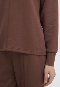Pullover marrone con una texture morbida, vestibilità rilassata, dettagli delle cuciture e un piccolo etichetta circolare sul bordo. Abbinato a pantaloni coordinati.