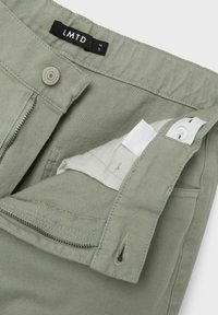 Pantaloni verde oliva chiaro con zip e chiusura a bottone. Presentano una texture liscia e una piccola tasca all'interno della cintura.