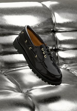 Chaussure en cuir verni noir avec œillets et lacets dorés, présentant une semelle noire à nervures audacieuses. Positionnée au-dessus d'une surface métallique brillante.