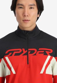Maglia sportiva zip-up rossa e nera con il logo "SPYDER" audace, colletto alto e dettagli e pannelli bianchi a contrasto. Tessuto morbido.