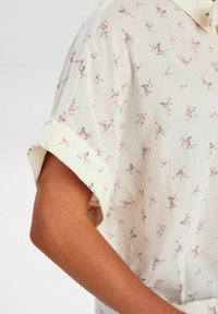 Floral short-sleeve blouse in cream fabric featuring pink flowers and green leaves, with a soft texture and rolled sleeve cuffs.