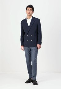 Marineblauer, doppelt geknöpfter Blazer mit zwei Fronttaschen, kombiniert mit strukturierten grauen Hosen und schwarzen Schuhen. Darunter ein weißes Hemd mit Kragen.