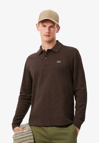 Polo a maniche lunghe marrone con logo ricamato verde, abbinato a un cappellino beige e che tiene un tessuto a righe grigie e bianche.