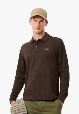 Polo a maniche lunghe marrone con logo ricamato verde, abbinato a un cappellino beige e che tiene un tessuto a righe grigie e bianche.