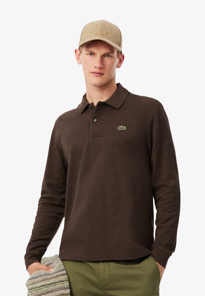 Polo a maniche lunghe marrone con logo ricamato verde, abbinato a un cappellino beige e che tiene un tessuto a righe grigie e bianche.