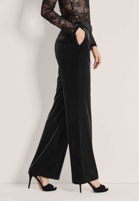 Zwarte fluwelen wijde pijpen broek met een relaxte fit, voorzien van zijzakken en een gladde, zachte textuur. Gecombineerd met hoge zwarte schoenen.