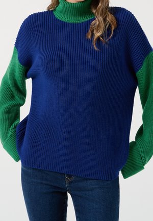 Femme portant un pull en maille côtelée avec col roulé et manches vertes, torse bleu, associé à un jean bleu foncé.