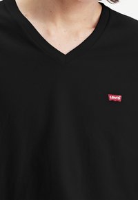 Camiseta negra de cuello en V, hecha de material suave, con un pequeño parche del logo rojo de Levi's en el lado izquierdo del pecho. Textura suave.