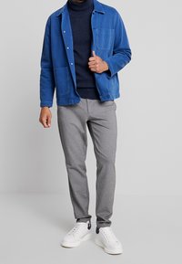 Veste en denim bleu portée sur un pull marine, pantalon gris fuselé, et baskets blanches avec des accents foncés. La veste comprend deux poches avant.