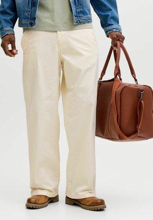 Mężczyzna w jasnożółtych spodniach, brązowych butach i dżinsowej kurtce, trzymający dużą brązową torbę podróżną ze skóry za uchwyty.