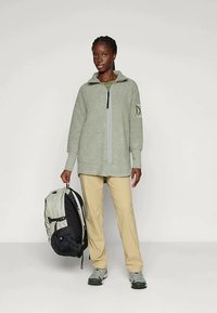 Gaiši zaļš rāvējslēdzēja jaca ar teksturētu audumu, brīvs piegriezums, sānu kabatas, valkāta virs tumši zaļas blūzes un kopā ar bežīgiem biksēm un mugursomu.