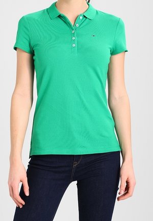 Frau, die ein eng anliegendes grünes Polo-Shirt mit kurzen Ärmeln und Knöpfen sowie dunkelblaue Jeans trägt und vor einem einfarbigen Hintergrund steht.
