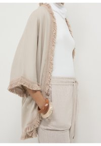 Beige kimono-stijl wrap met franje-oevers, gemaakt van zachte stof. Het model draagt een witte coltrui en lichte broek, en houdt houten armbanden vast.