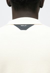Πόλο μπλούζα σε εκρού χρώμα με υφή πλεκτού. Διαθέτει μαύρη τζην λεπτομέρεια στη βάση του λαιμού με την επιγραφή "REPLAY" σε λευκό.