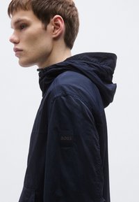 Tmavomodrá kapucňová bunda z ľahkej tkaniny, s nášivkou loga na ľavom rukáve a voľným strihom.