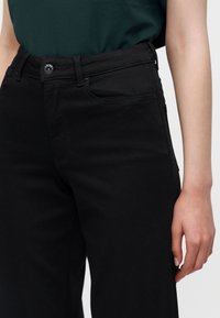 Femme portant un jean noir taille haute avec poches devant et fermeture par bouton, associé à un haut vert foncé rentré.