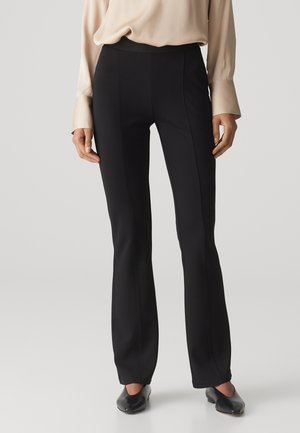 Vrouw draagt zwarte, nauwsluitende broek met voornaad, beige blouse met lange mouwen en zwarte schoenen met lage hakken, tegen een effen achtergrond.