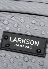 Tessuto grigio testurizzato con motivo a goccia d'acqua in rilievo. Presenta un'etichetta nera con testo bianco embossato "LARKSON HAMBURG" e accenti lucidi.